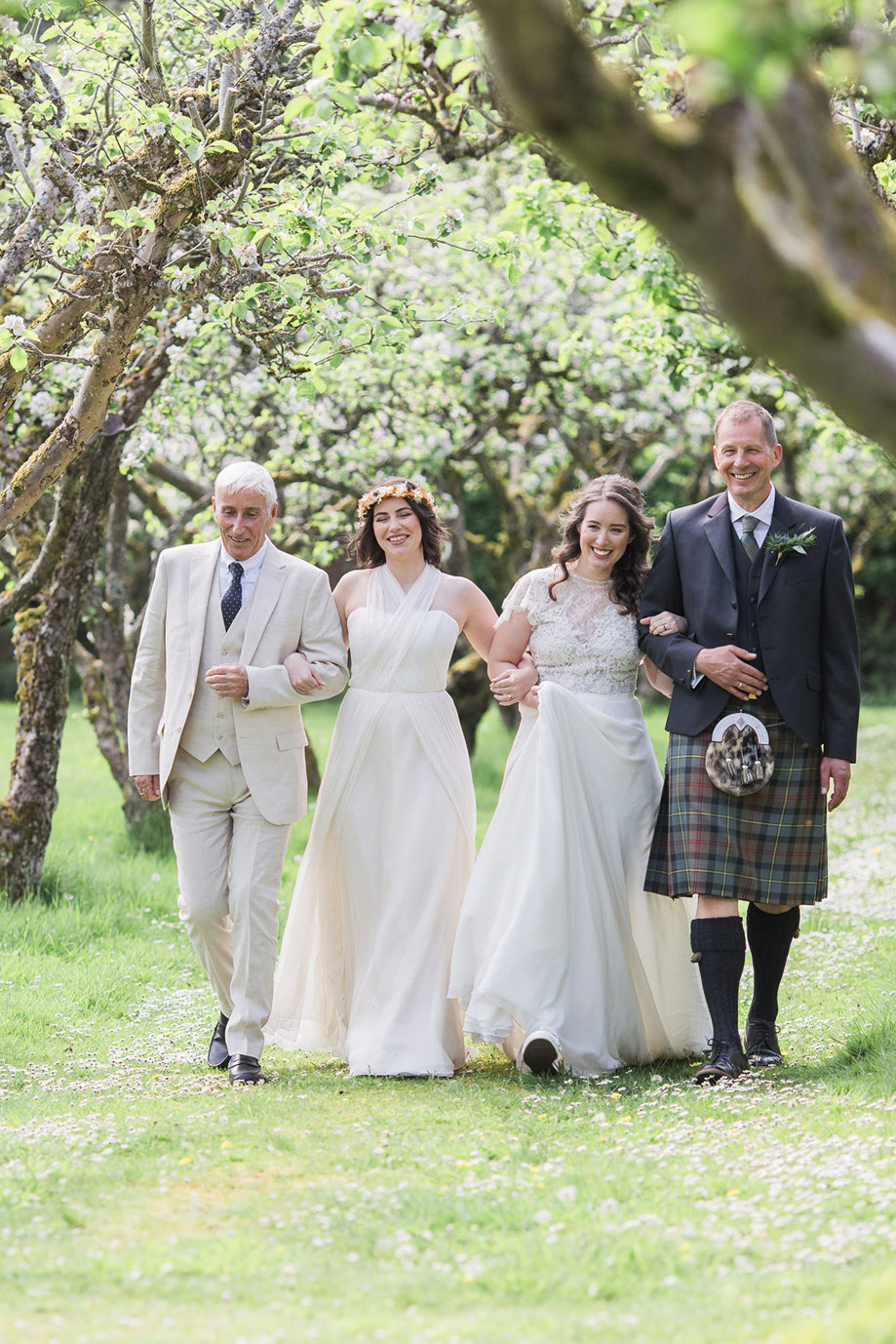 a row of four people walking across grass under blossoming trees with their arms interlinked