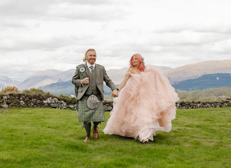 A man wearing a light green kilt and a woman wearing a pink wedding dress walking through a field with hills in the background