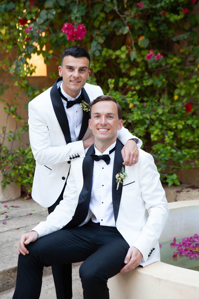 two smiling wearing white suit jackets and bow ties grooms posing in front of a green hedge with dark pink flowers