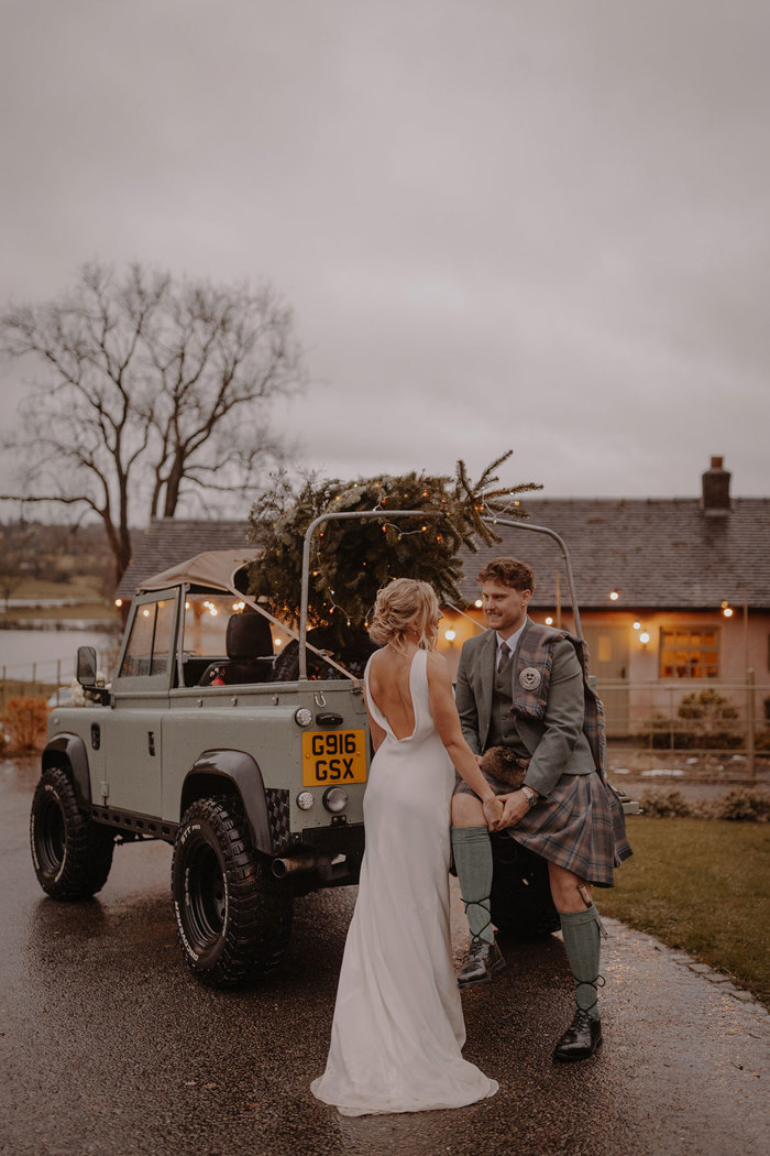 blonde bride in open back white wedding dress faces groom in green jacket and red, blue, green kilt with matching fly plaid who is sitting along the back of green truck with Christmas tree hoisted up in the middle