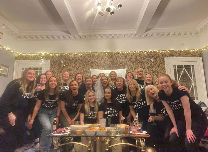 Group of women wearing black tshirts with a woman standing in a white tshirt and a veil in the middle, behind a table of food and drinks