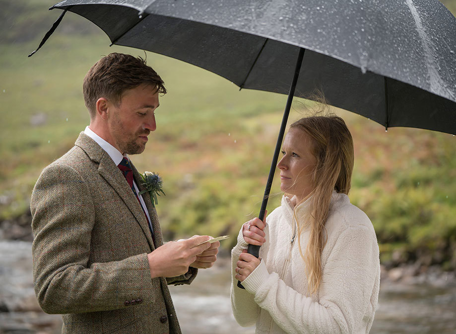 a groom reads from a small piece of paper to a bride wearing a cream fleece. They stand under a large umbrella. There is a river and a heather covered hill in the background