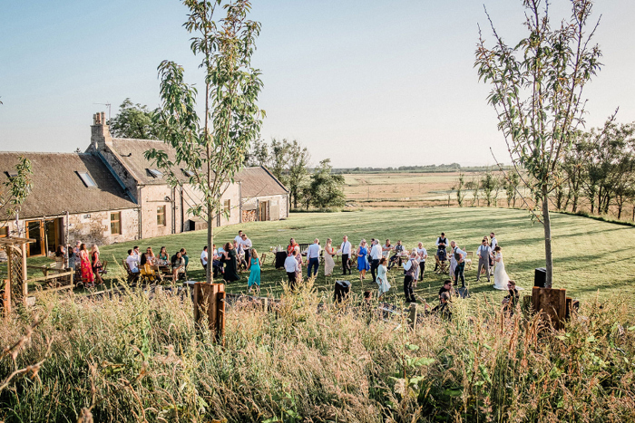 people gathered on grass outside of a small stone building 