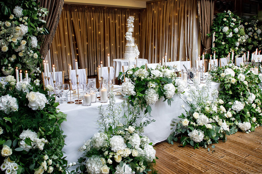 a long table decorated for a wedding with abundant white flowers, green foliage and white candles.