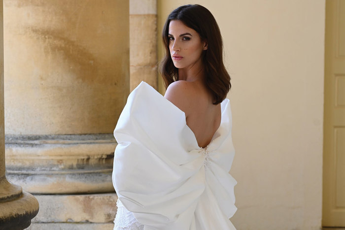 a model wearing a large oversize bow wedding dress looking over her shoulder standing in a pale yellow stone building