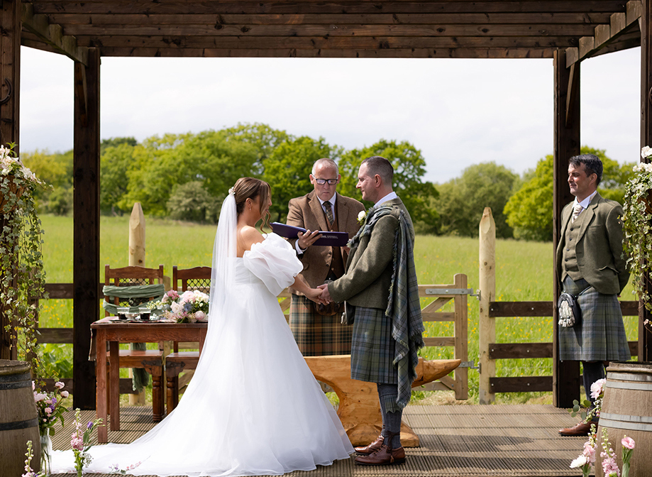 bride and groom facing one another holding hands surrounded by two men and fields and flowers