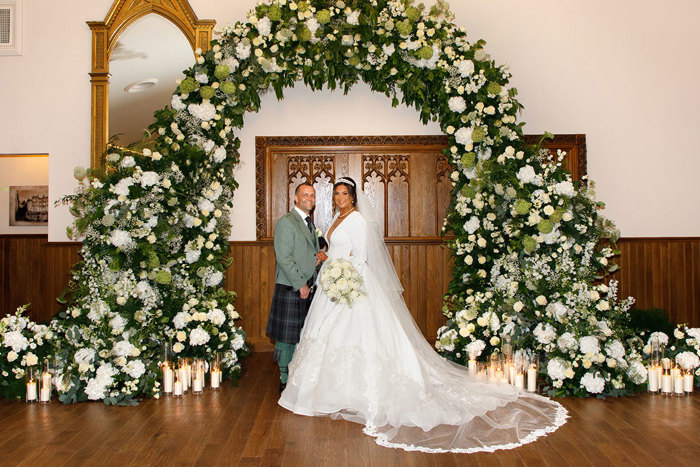 a bride and groom standing under a large arch of white flowers and green foliage at Crossbasket Castle.