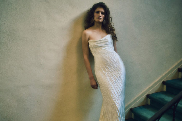 a model wearing a crinkled silk strapless wedding gown standing on stairs leaning against a wall