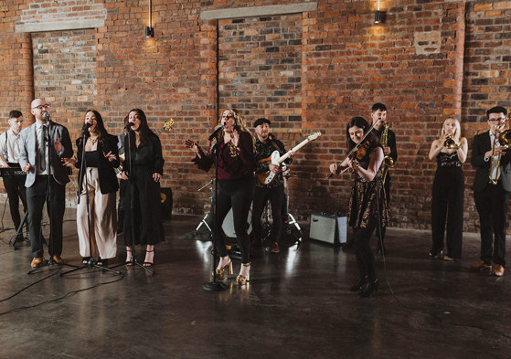 a large group of musicians each playing a different instrument together stand in a large unfurnished room with a large brick wall behind them