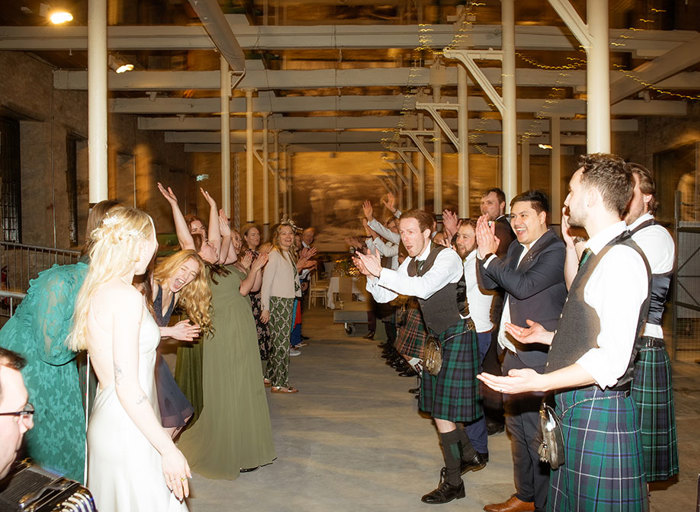 two rows of people lined up to do a Scottish ceilidh dance at a wedding in a room with exposed metal beams