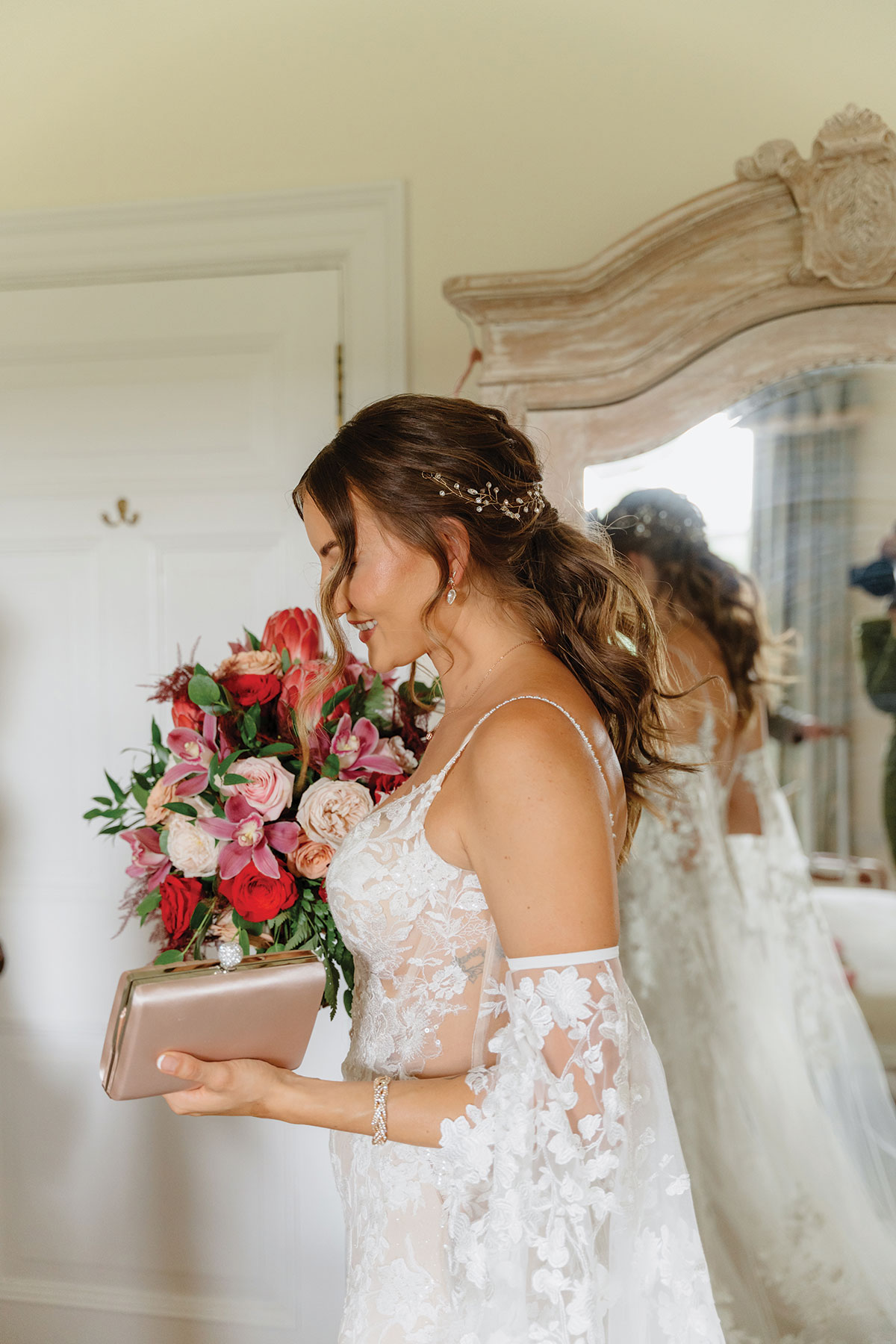 side view of bride in floaty floral embroiderd corset bodice wedding dress stood in front of a red and pink flower display and a large wardrobe mirror