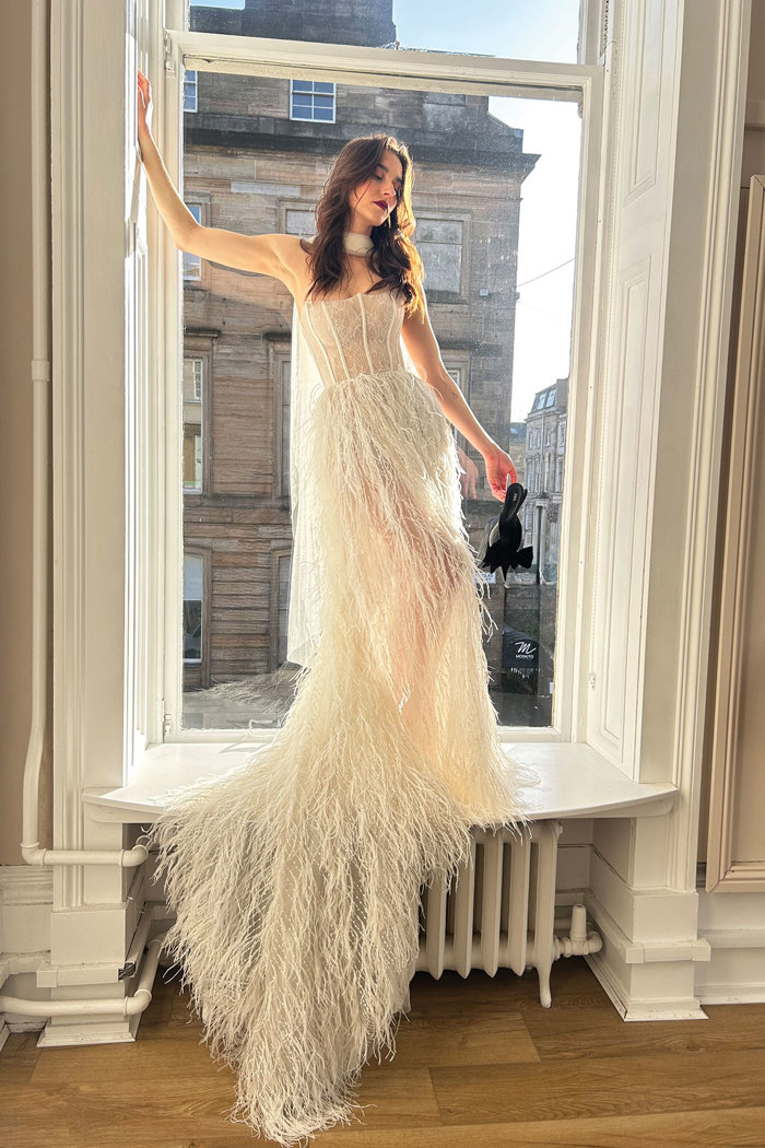 a bride wearing a feather wedding dress standing in a window. Fleur by Wona Concept.