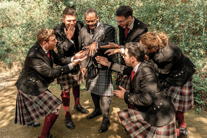 groom stands surrounded by five groomsmen, all wearing tartan kilts, as they all gesture in admiration and surprise to the groom's wedding ring