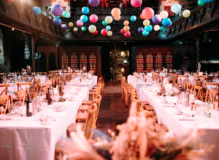 long tables set for a wedding dinner flanked by wooden crossback chairs below rows of colourful hanging lanterns