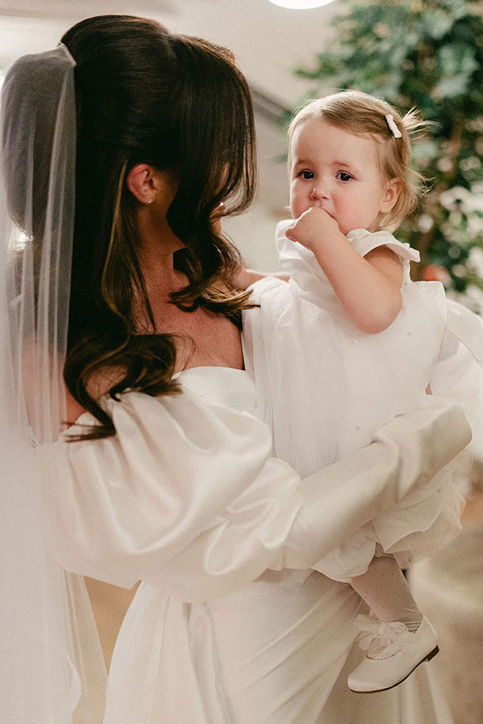 brunette bride in veil and wedding dress with puff sleeves holds toddler in white flowergirl dress