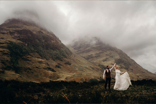 A bride and groom dancing together on a hilltop with other hills covered in mist in the background