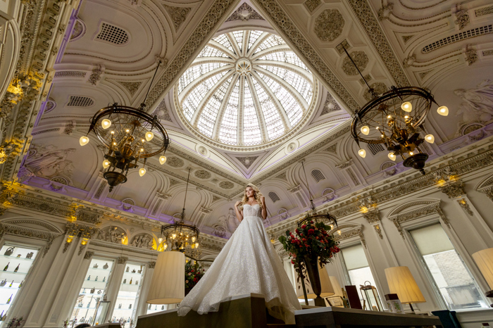 A bride in a long dress standing on a table with the camera pointing upwards towards her showing an ornate dome ceiling above her