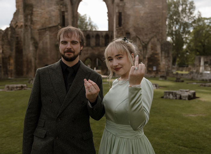 A bride and groom standing outside Elgin Cathedral, both holding up their hands to display wedding rings