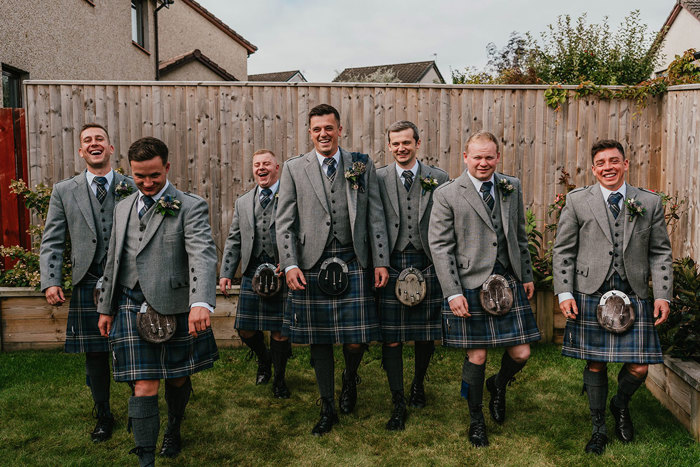 Seven men in grey and blue kilts walk across grass in a garden 