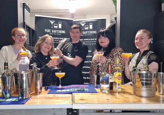 Four women holding glasses with cocktails surrounding one man holding cocktail shaker