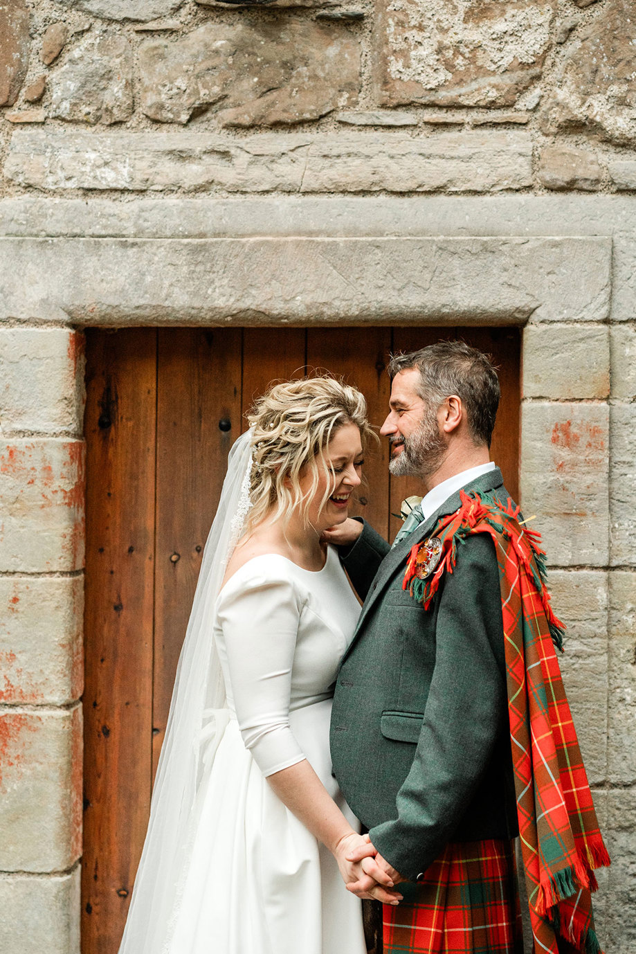 bride in white wedding dress and long veil and groom in red and green tartan kilt have their fingers intertwined as they stand facing each other outside a wooden door in a grey stone building