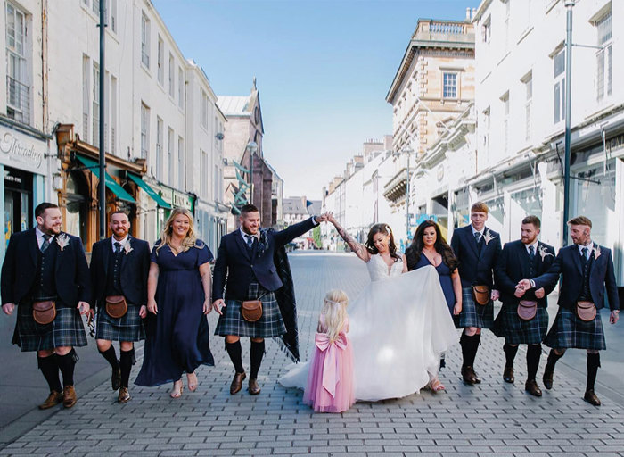 A groom and five groomsmen wearing matching blue and green kilts walk down a street with a bride in a white dress, two bridesmaids in navy dresses and a flower girl in a pink dress