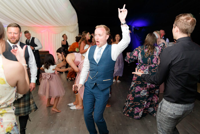 People on a dance floor with a man in a blue suit as the focus with one hand holding a drink and the other in the air