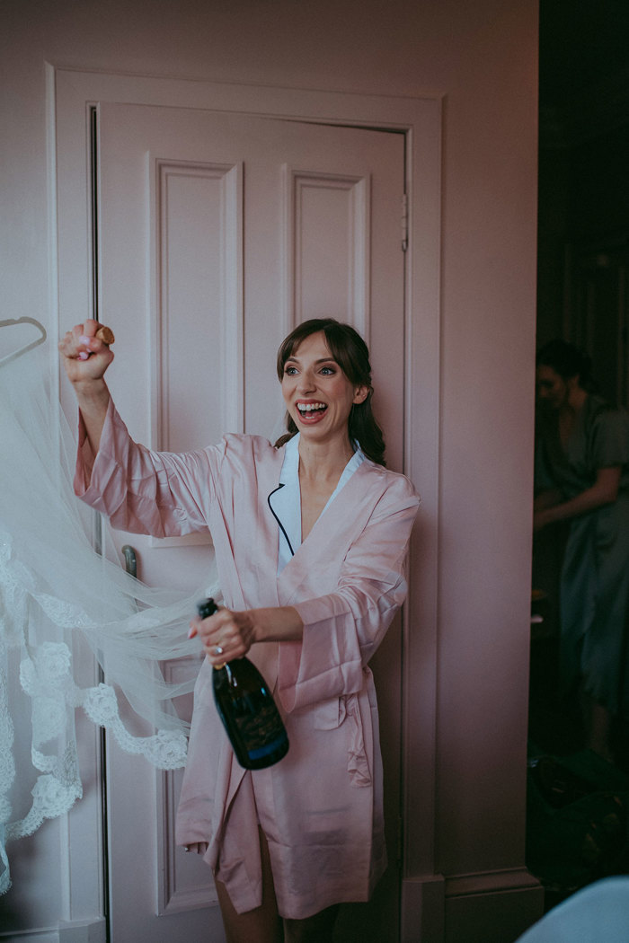 a person wearing a pale pink robe and white pyjamas opening a bottle of champagne in front of a pink door.