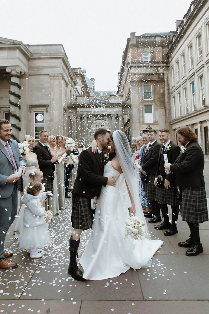 A bride and groom standing outside surrounded by their wedding guests as white confetti is thrown over them