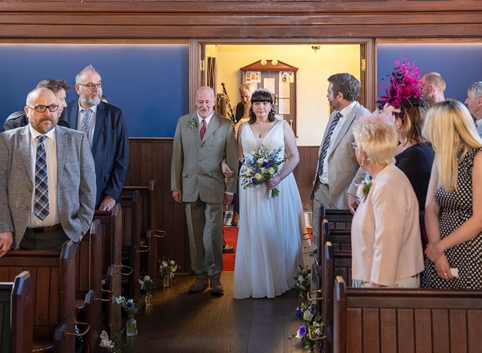 a pensive looking bride walks down the aisle on the arm of a man wearing a suit. Rows of wedding guests standing at wooden pews turn to watch her enter.