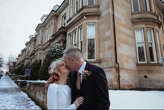 a man and woman kiss in front of yellow sandstone building with snow on the ground 