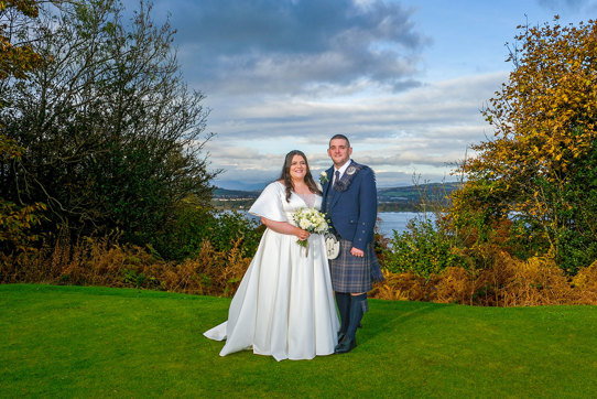 A bride and groom stand next to each other facing the camera with a view of hills and water behind them