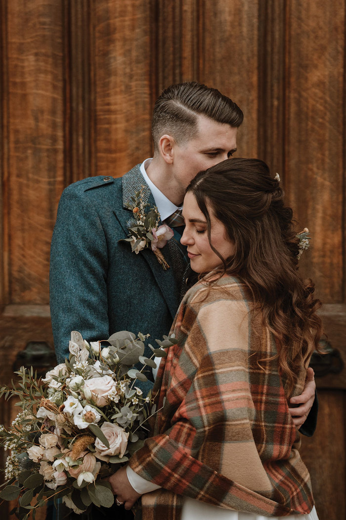 bride holding a pale neutral bouquet and draped in a brown tartan shawl looks down as groom nestles his head into her hair