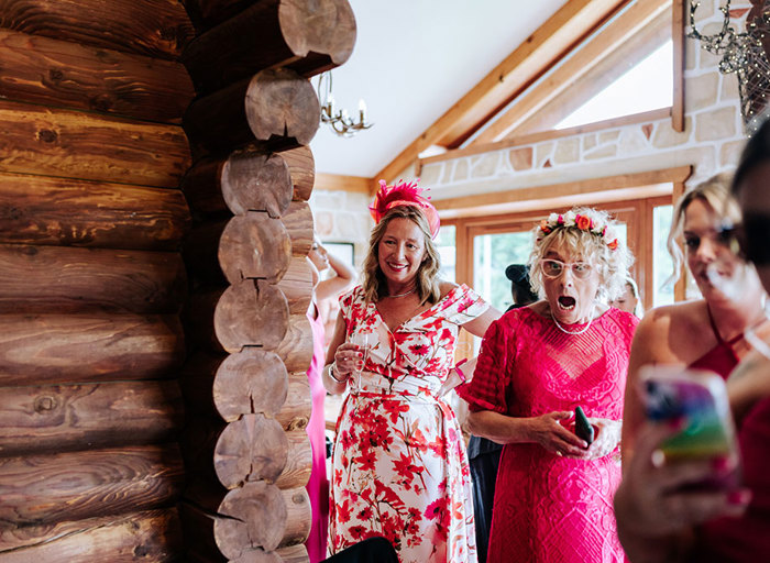 a lady wearing a floral dress and a shocked looking older lady wearing a hot pink dress and flower crown in a wooden building setting