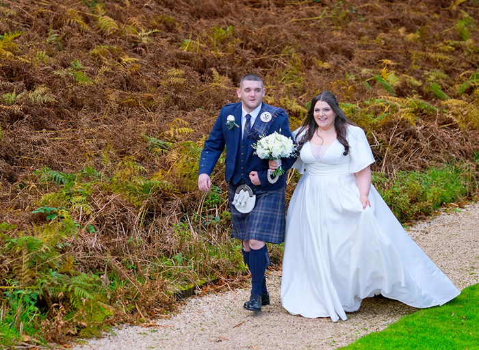 a bride and groom walk along a path holding hands in a woodland area