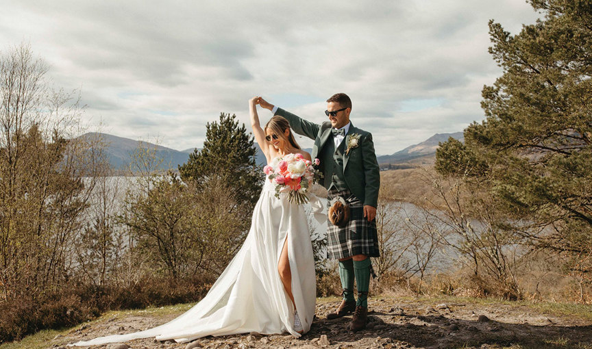 A bride dances under a groom's arm. The bride is holding a large pink and white bouquet of flowers and wearing a textured wedding dress with skirt split. The groom is wearing a green tweed kilt outfit with bow tie. They are standing on stony ground with trees, hills and Loch Lomond in the background