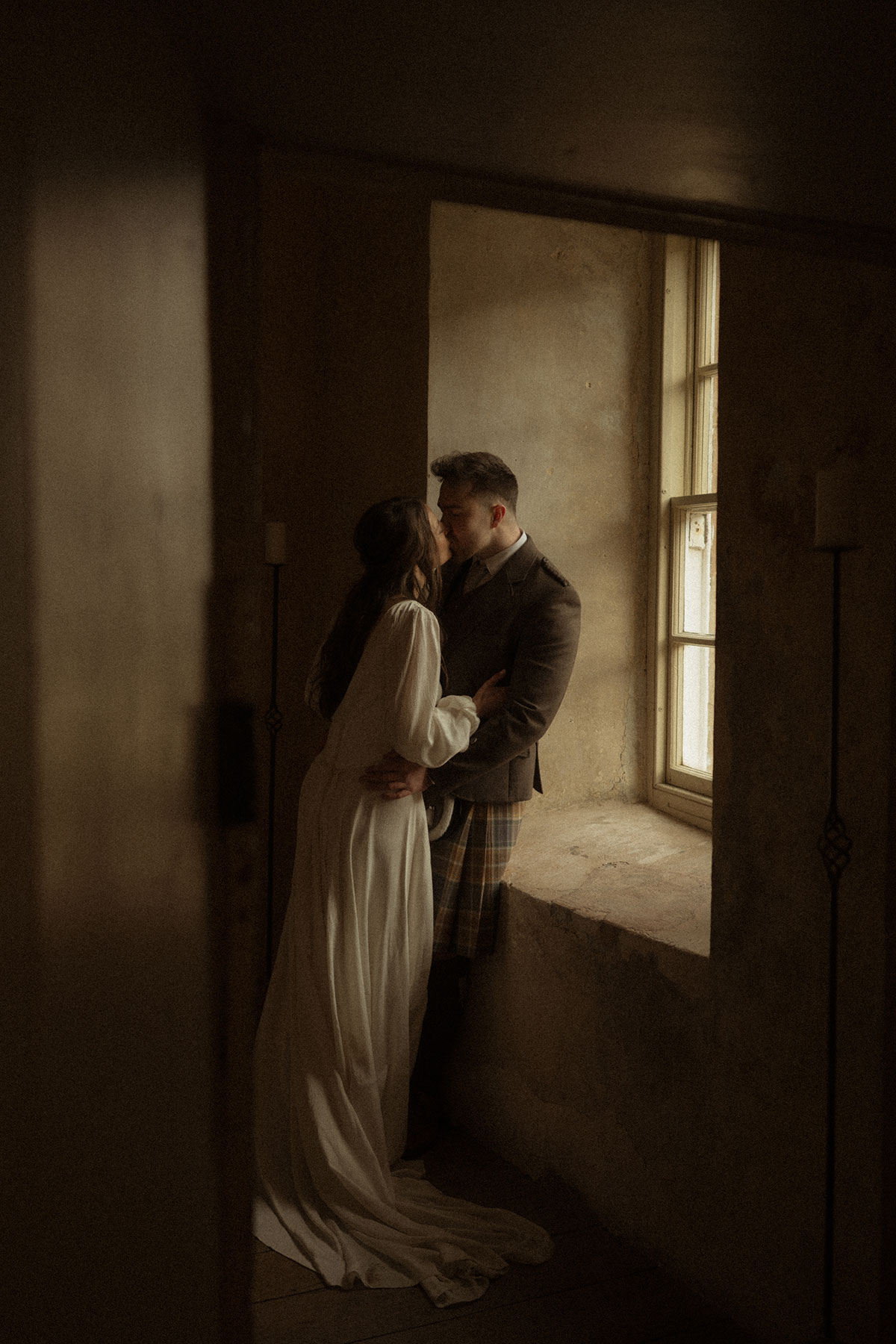 bride in trailing satin dress leans in, kissing groom in kilt outfit, leaning against stone windowsill in a shadowy room