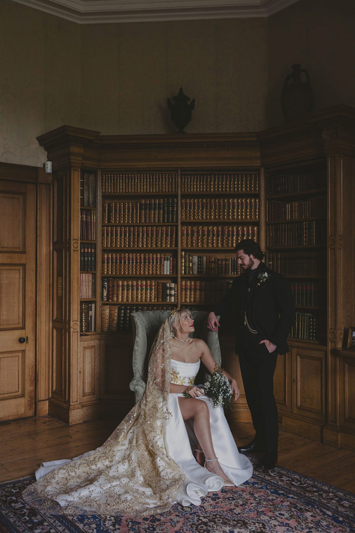 A groom in a suit and a bride in a white dress sitting in a chair. They are in front of a bookcase filled with books.