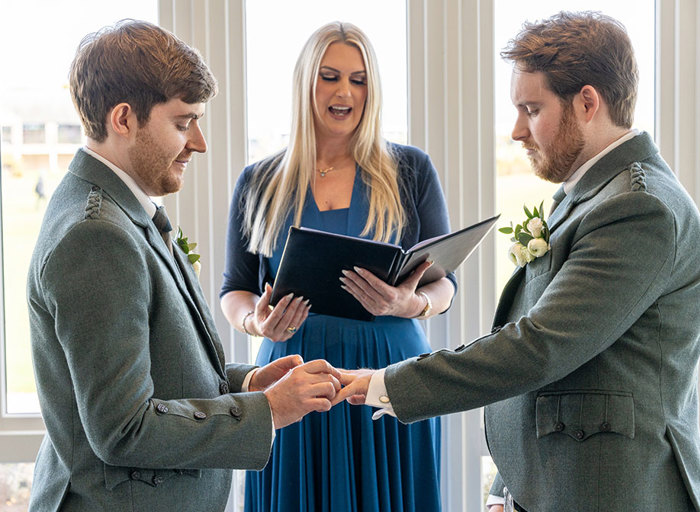 two grooms wearing green tweed kilt outfits exchange wedding rings in front of a celebrant