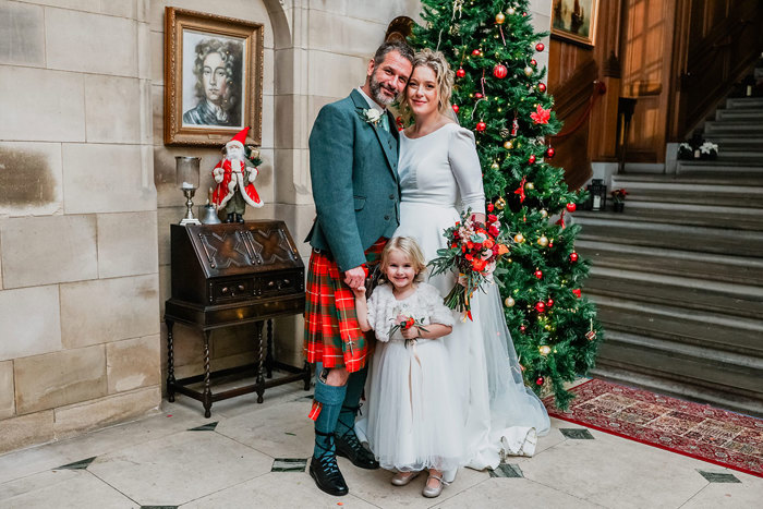 bride in white wedding dress and veil and groom in red and green tartan kilt stand in front of Christmas tree with a young flower girl dressed in a little white dress stood between them