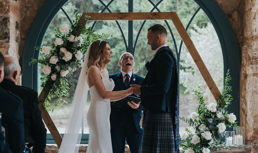 a bride and groom standing in front of a celebrant and an arched window during a wedding ceremony.