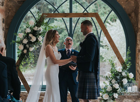 a bride and groom standing in front of a celebrant and an arched window during a wedding ceremony.