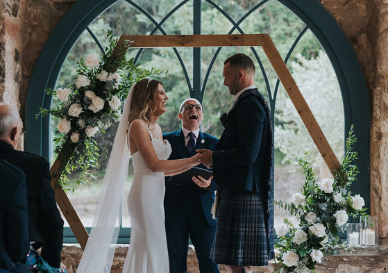 a bride and groom standing in front of a celebrant and an arched window during a wedding ceremony.