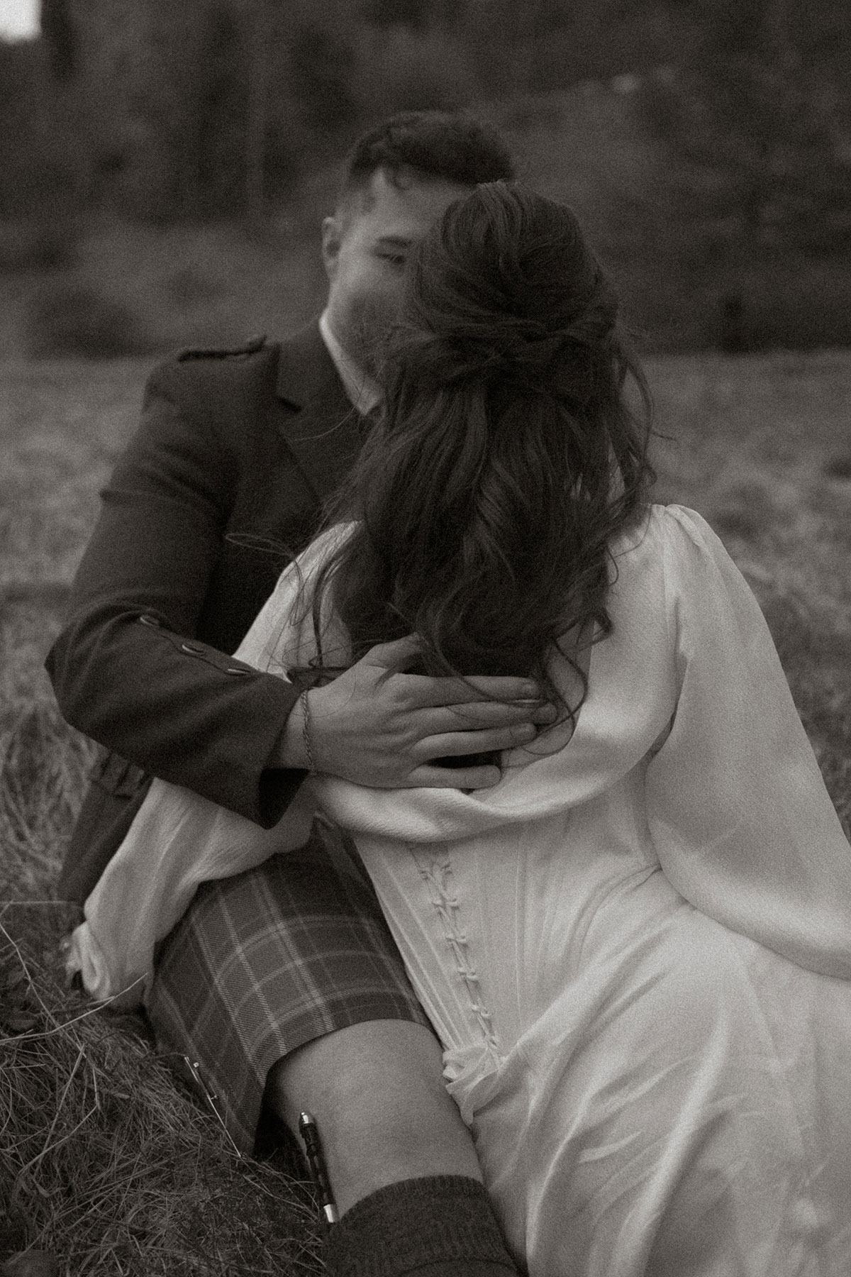 black and white photo of dark haired bride in a corset dress, with her back to the camera, facing groom in kilt outfit who has a hand softly in the middle of her back