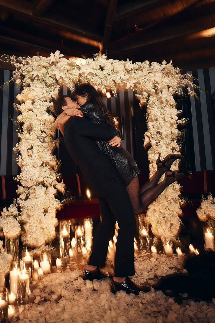 A man lifting a woman in an embrace as they kiss in front of a white flower arch surrounded by candles and flower petals