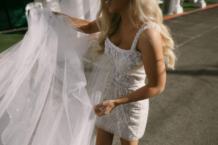 bride in embellished white mini dress holds her long veil out in front of herself to adjust