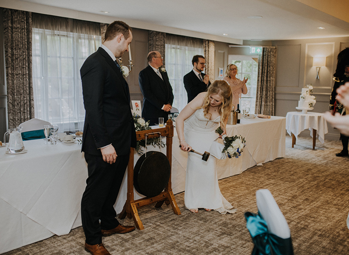 A bride and groom stand in front of a long table with a white table cloth and the bride hits a gong with a hammer