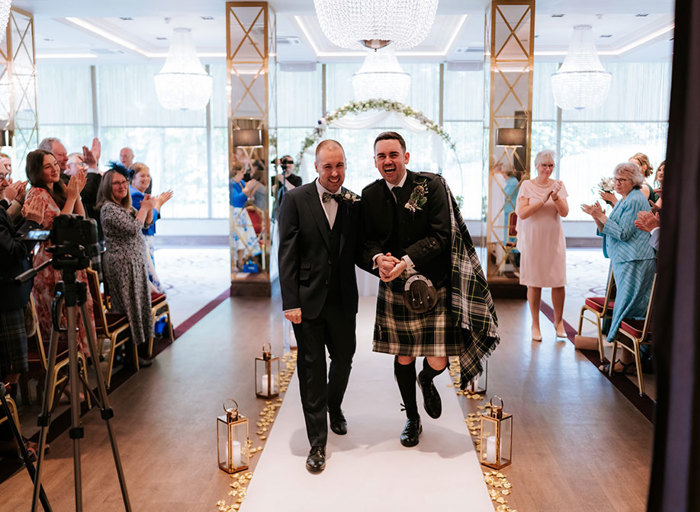 two elated-looking grooms holding hands walking up a white carpet aisle as rows of wedding guests either side clap and cheer