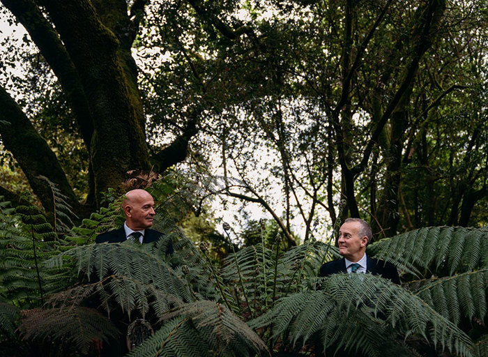 two men standing behind huge fern leaves below trees