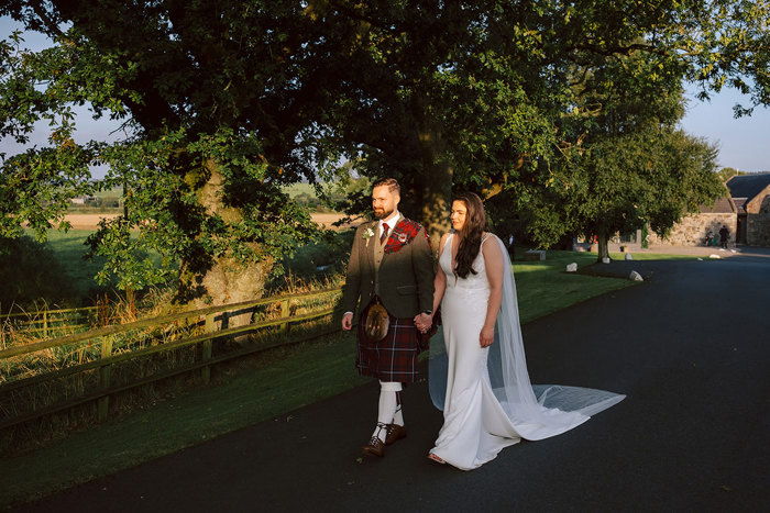 A bride and groom walk holding hands through a farm with fields and trees behind them 